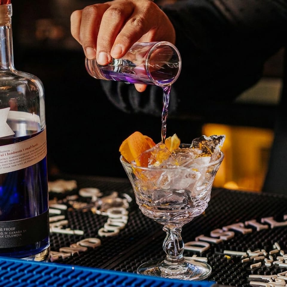 Bartender pours purple gin into an ice-filled stemmed glass garnished with citrus beside an Empress 1908 bottle.