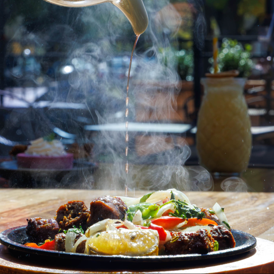 Steaming sizzling platter of grilled meat, vegetables, and lemon on a wooden table in a sunlit restaurant.