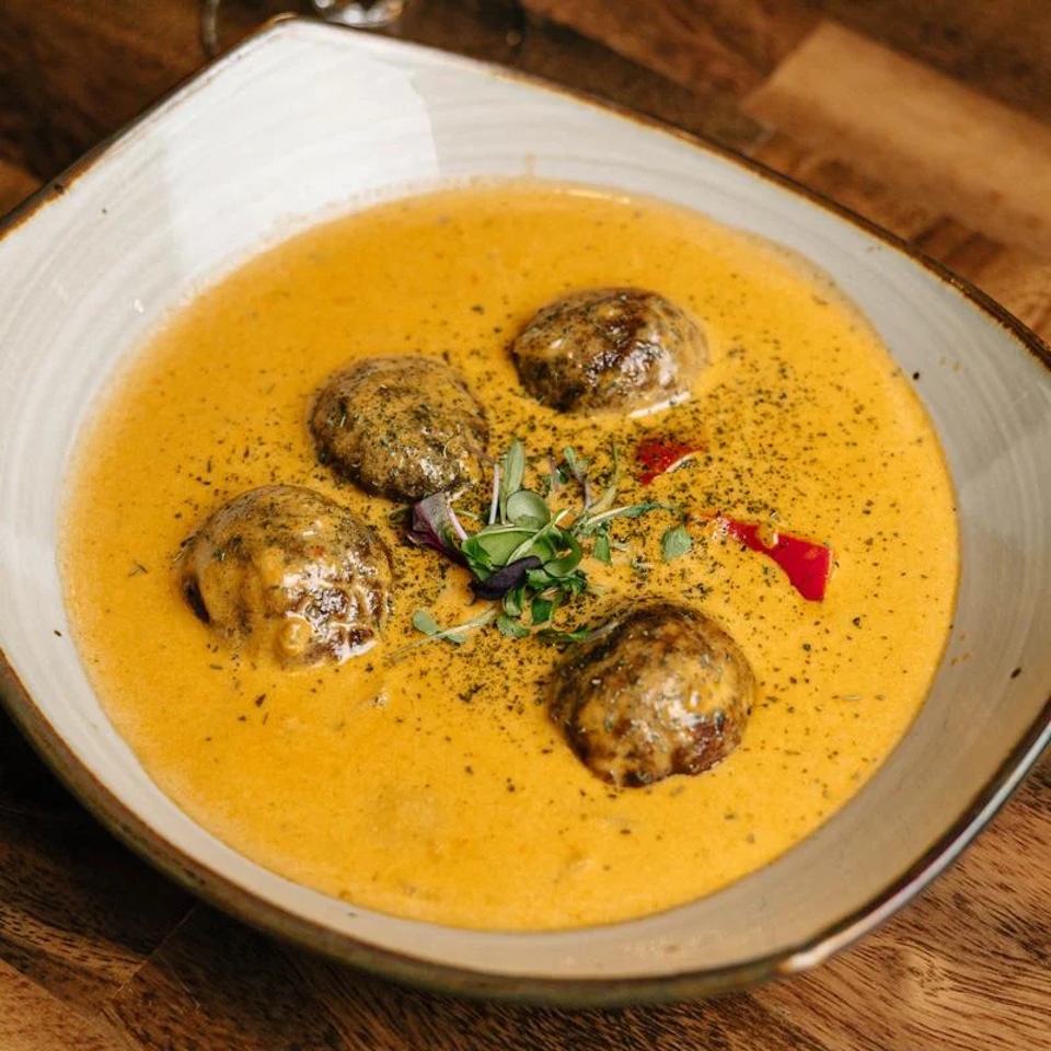 Four browned meatballs in creamy orange sauce, garnished with microgreens and red pepper, served in a shallow ceramic bowl.