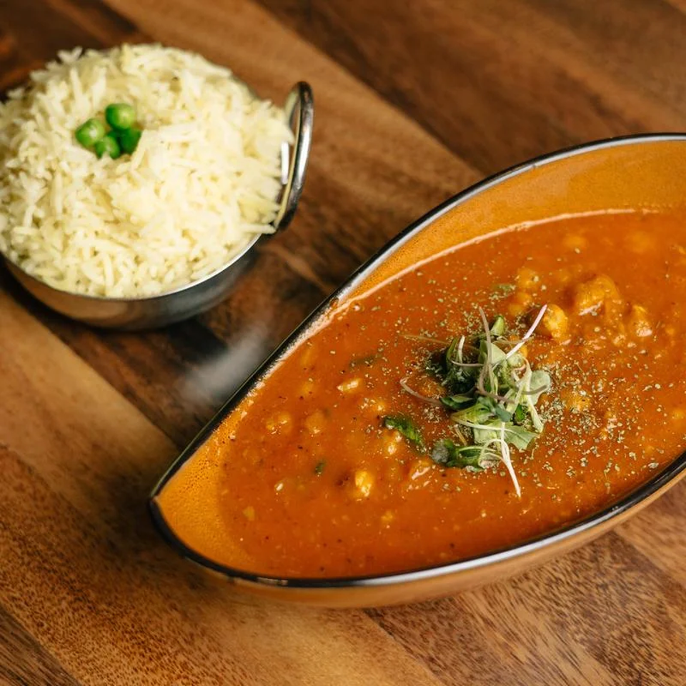 Bowl of creamy orange curry garnished with herbs, served alongside steamed white rice on a wooden table.