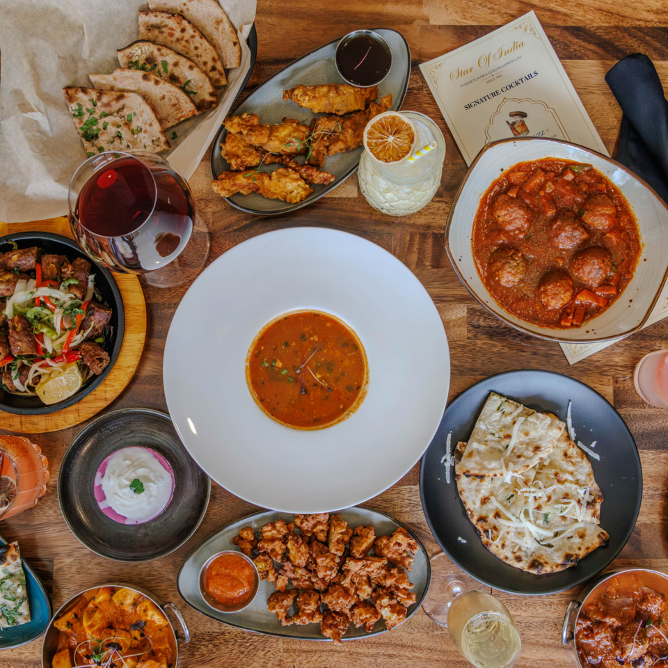 Overhead view of a wooden restaurant table filled with assorted dishes, flatbreads, grilled meats, curries, salads, and colorful drinks.