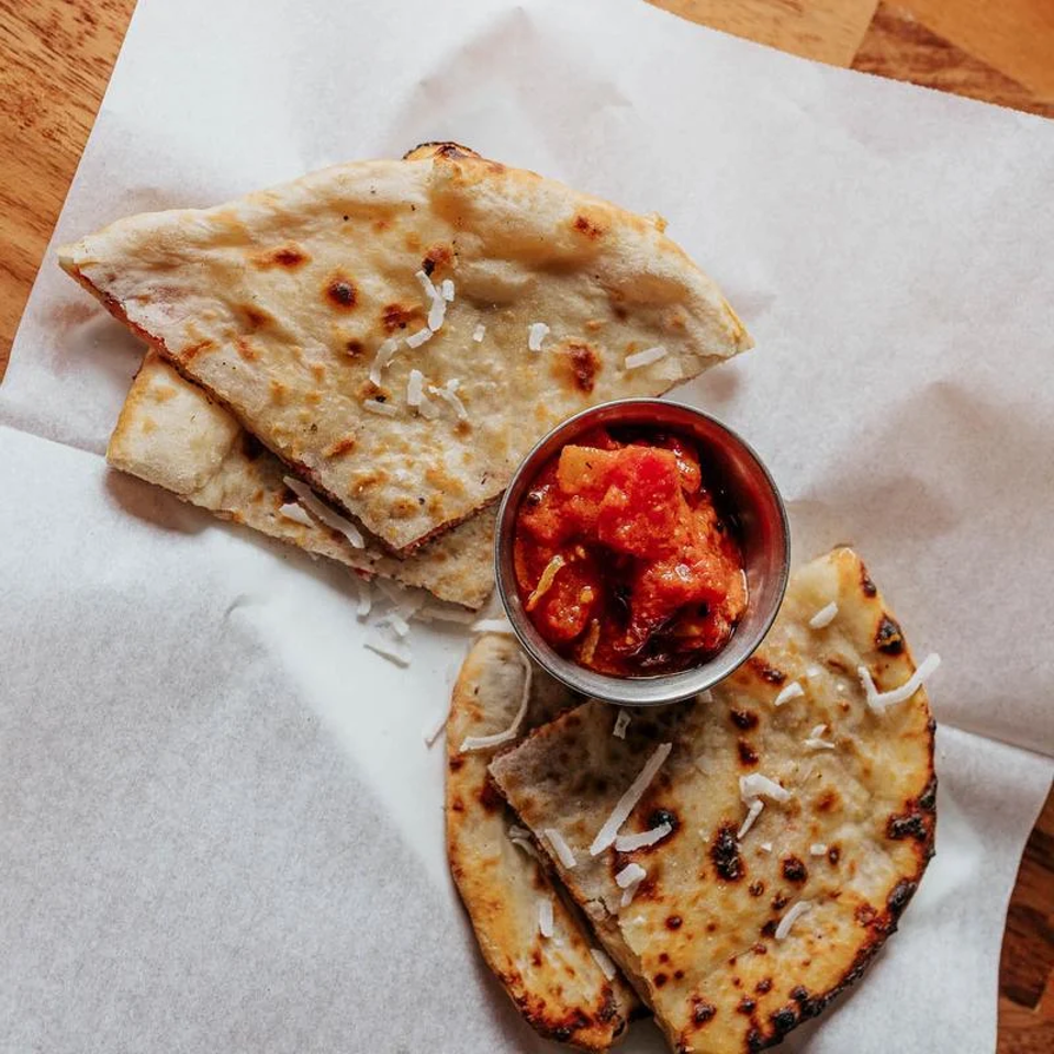 Grilled flatbreads sprinkled with shredded cheese surround a metal cup of chunky red tomato dipping sauce on parchment paper.