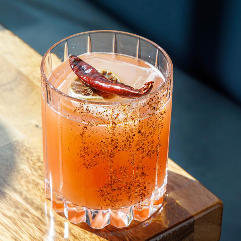 Orange-red cocktail in a ridged glass, garnished with a dried chili and lime, sits on a wooden table in sunlight.