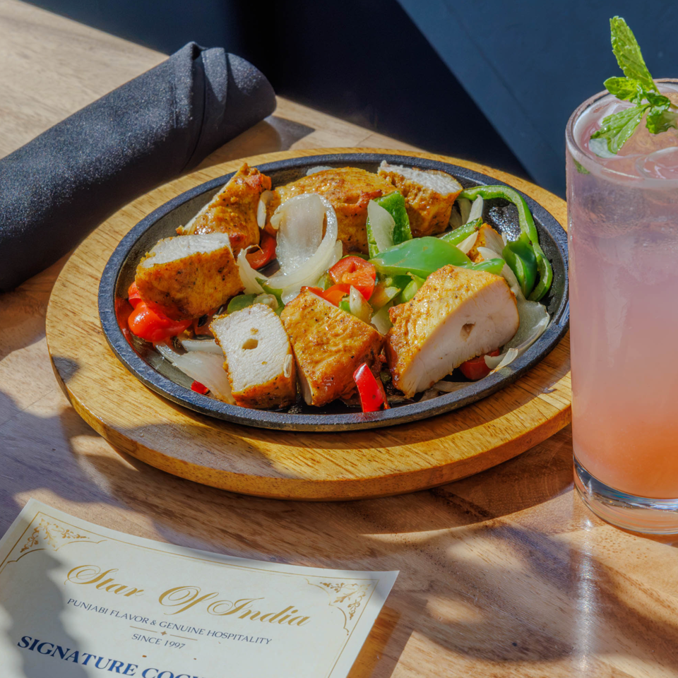 Indian grilled paneer and vegetables on a sizzling platter beside a pink mint-garnished drink and rolled napkin.