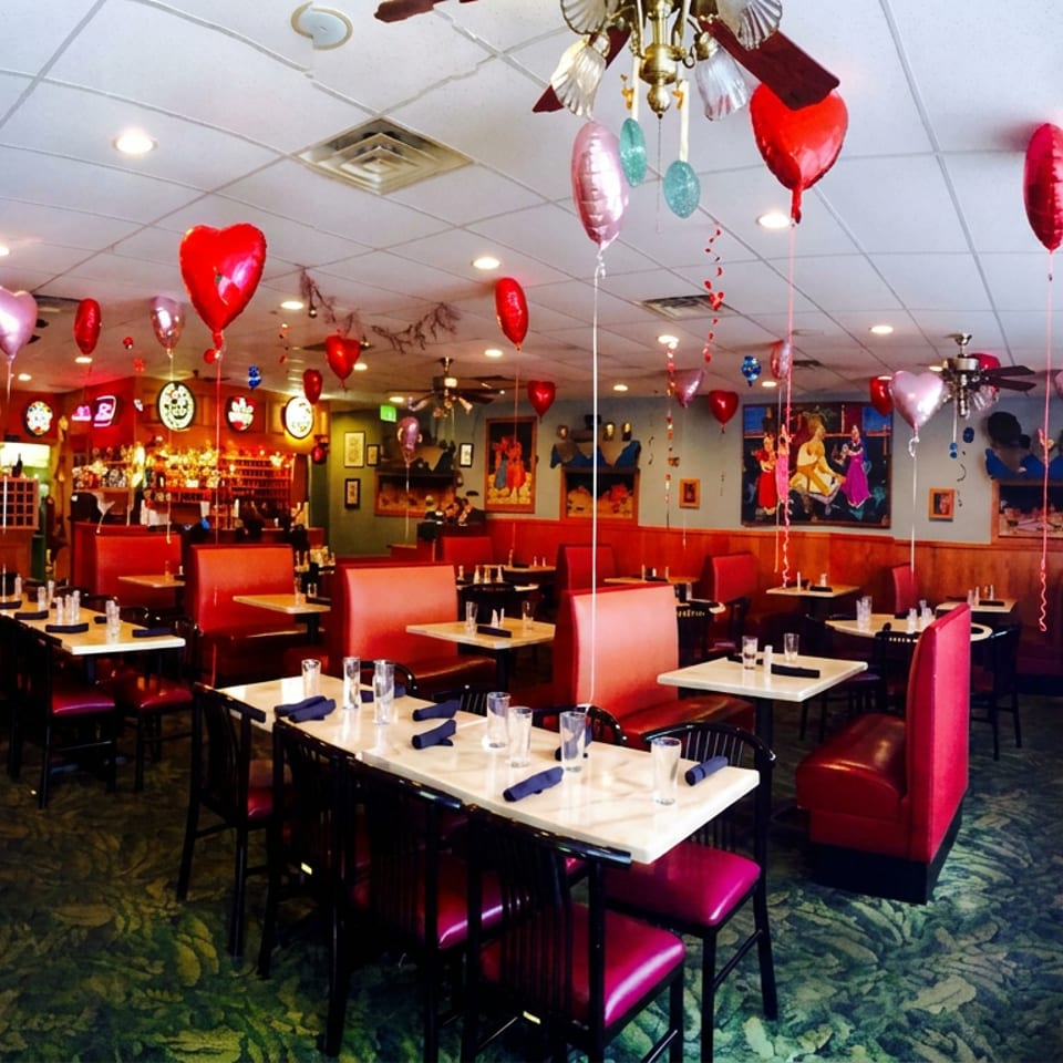 Festively decorated restaurant dining room with red booths, set tables, colorful heart-shaped balloons, and a visible bar.