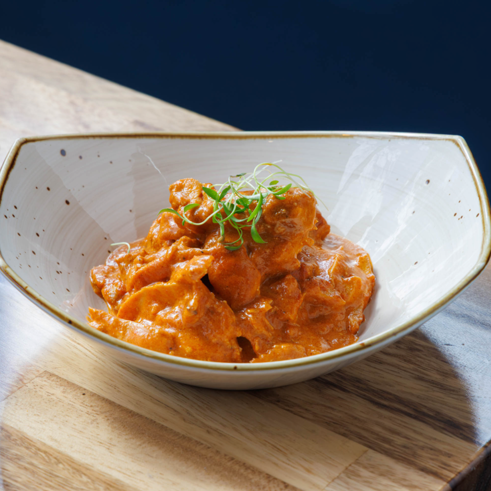 Bowl of creamy orange curry garnished with microgreens on a wooden serving board beside a folded black napkin.