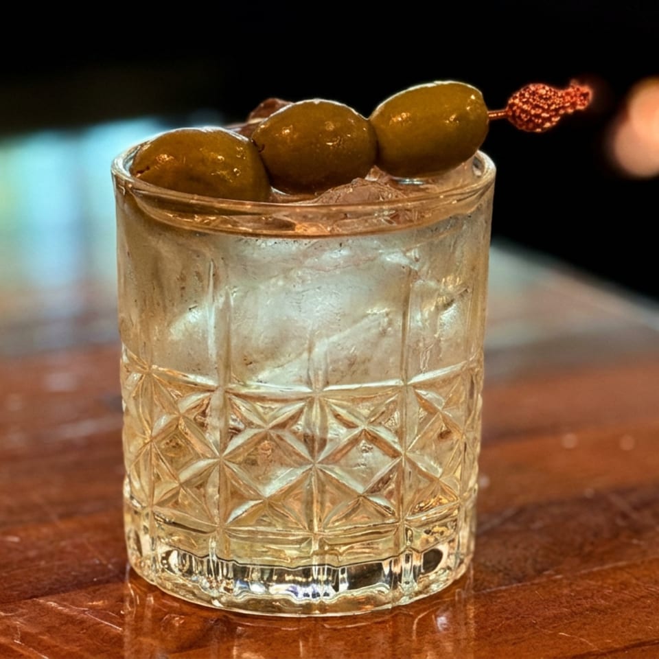 Cocktail in a textured rocks glass over ice, garnished with three green olives on a skewer, resting on a wooden bar.