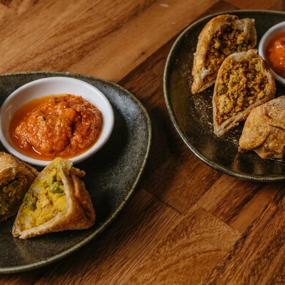 Two black plates hold assorted golden samosas and savory pastries with bowls of red dipping sauce on a wooden table.