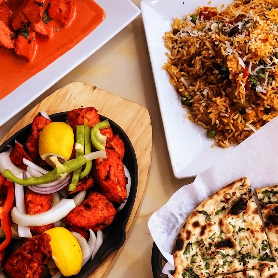 Indian meal featuring biryani, creamy red curry, tandoori kebabs with peppers and onions, and sliced naan.