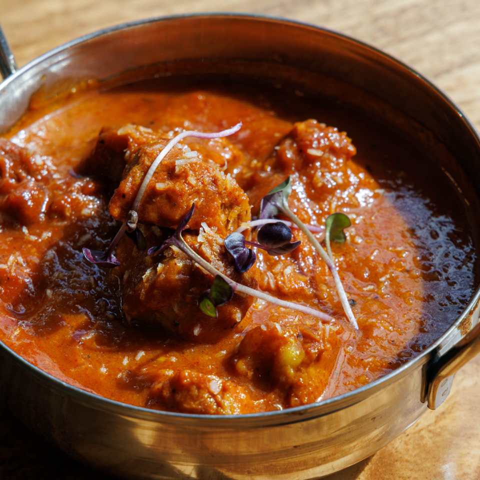 Copper serving pan filled with rich red curry, chunks of meat, and microgreens on a wooden table.