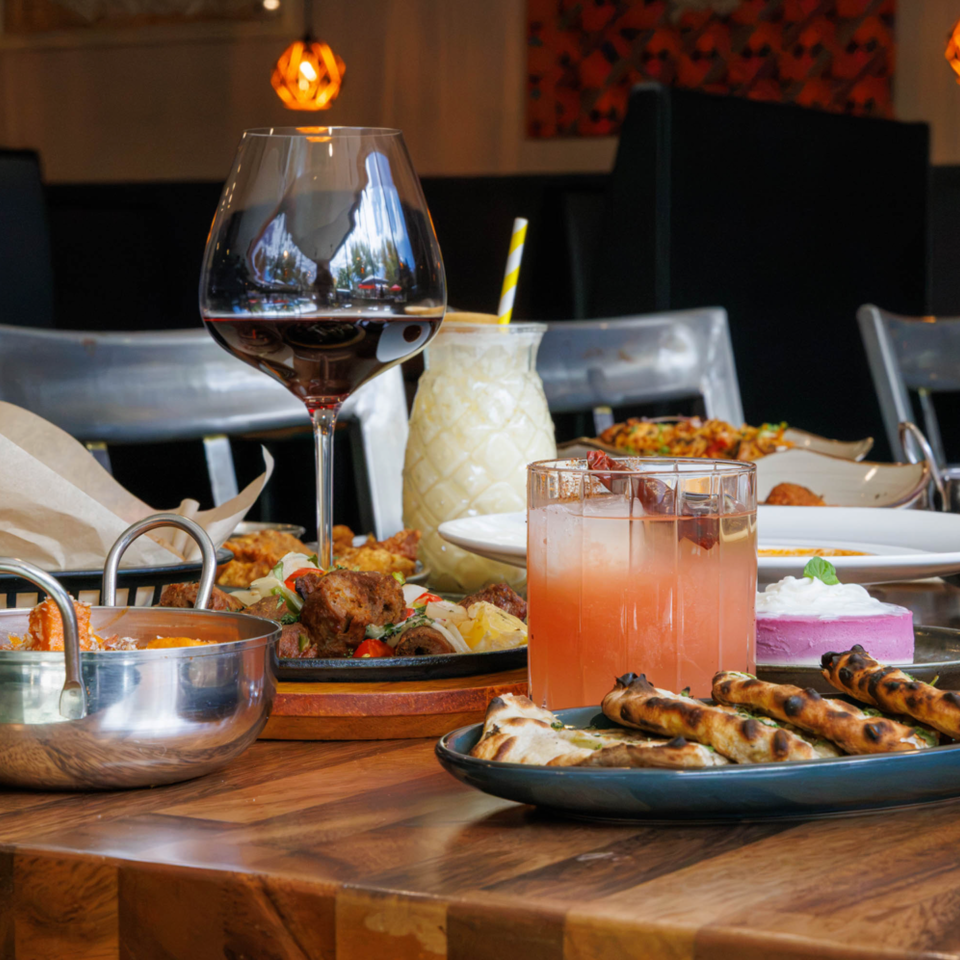 Restaurant table filled with Middle Eastern dishes, flatbreads, colorful desserts, cocktails, and a glass of red wine.