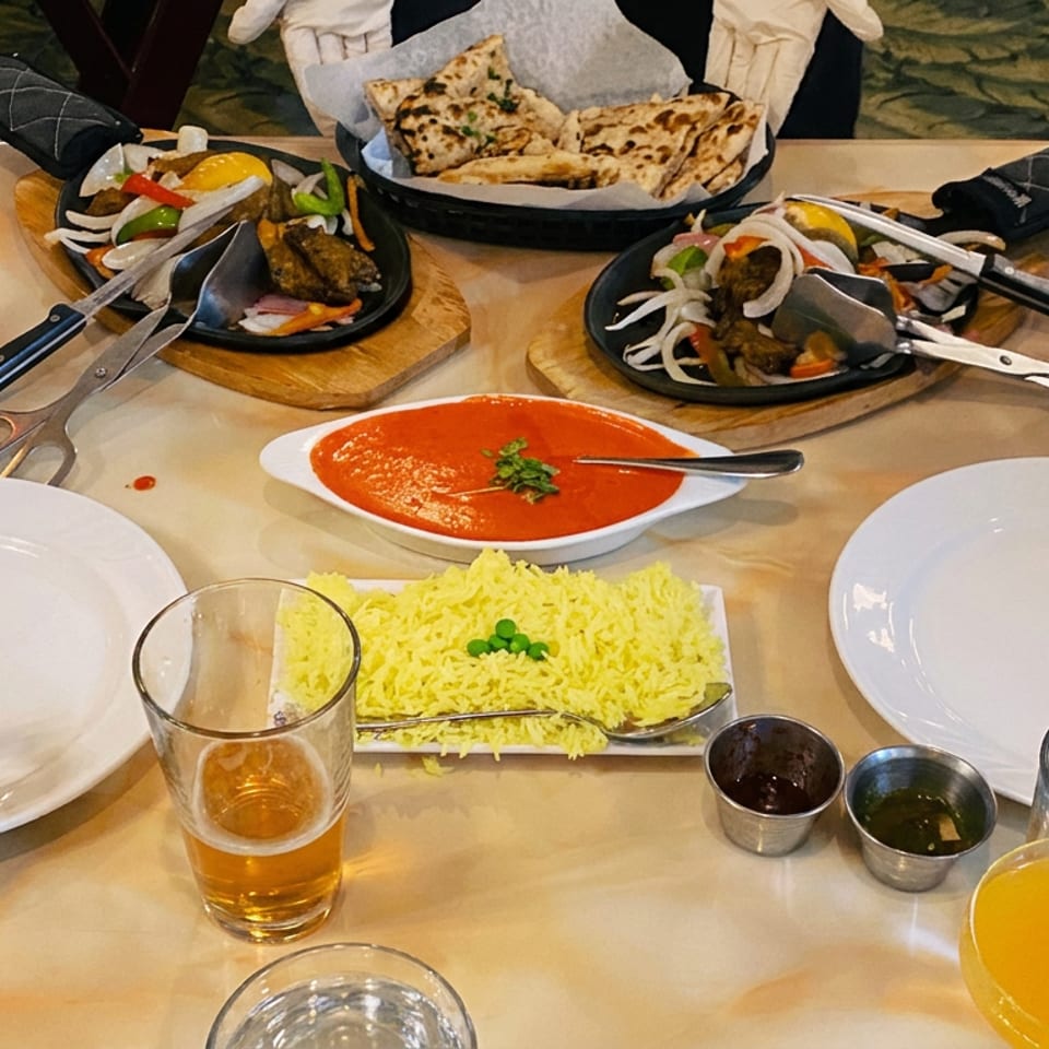 Restaurant table set with naan, yellow rice, red curry, grilled meats, vegetables, sauces, drinks, and diners’ hands.